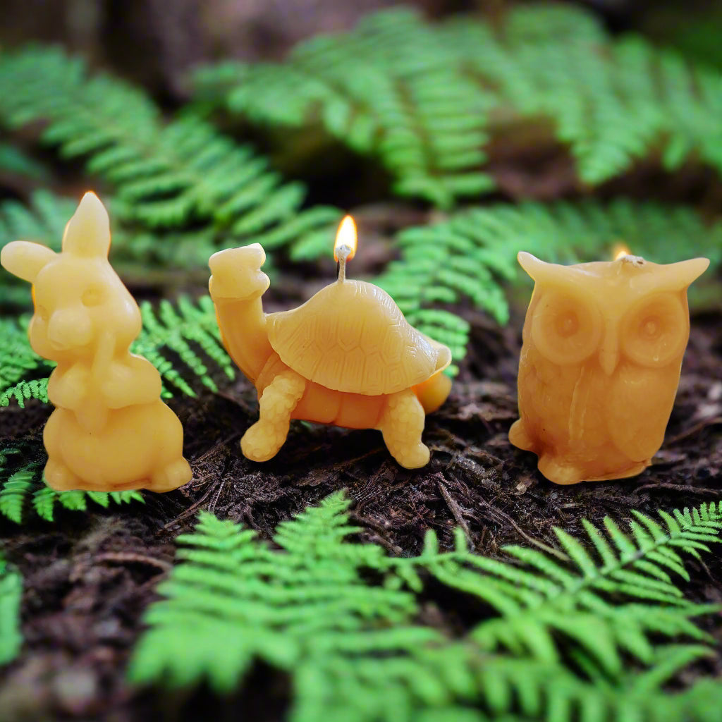 A set of three beeswax candles shaped like forest animals: a rabbit, a turtle, and an owl, displayed on a fabric with a floral pattern.