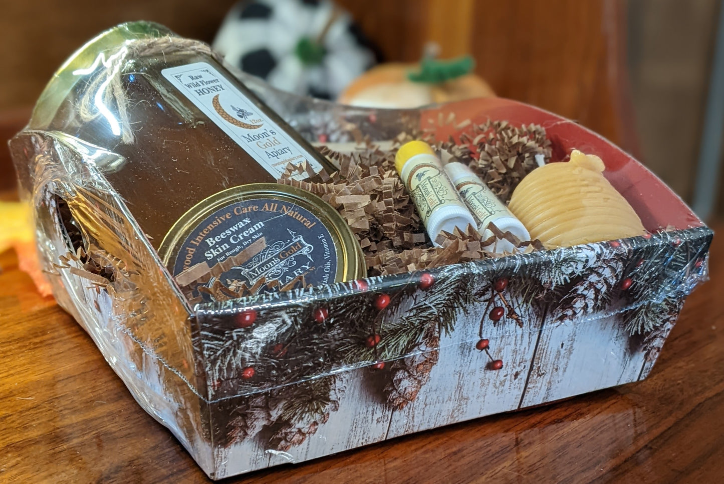 A small gift basket containing a jar of honey, skin cream, lip balms, hand-crafted soap, and a decorative skep hive candle, all presented in a festive wrapped arrangement.