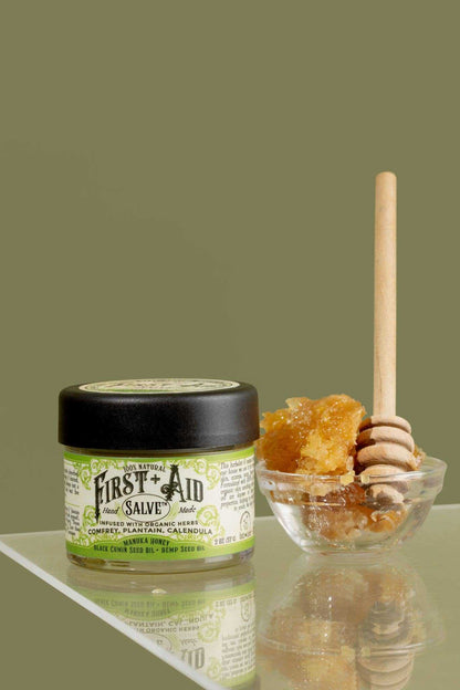 First Aid Salve in a clear glass jar with black lid and green botanical label, beside golden honey and wooden dipper