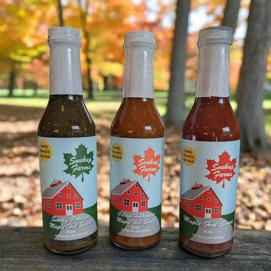 Hot sauce bottles with maple-leaf labels and red barn artwork, NY MAPLE PRODUCTS BY SOUKUP FARMS, in three shades.